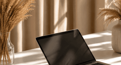 Modern laptop egy letisztult fehér íróasztalon, mellette elegáns üveg- és kerámiavázák száraz pampafűvel, lágy, napfényes árnyékokkal.